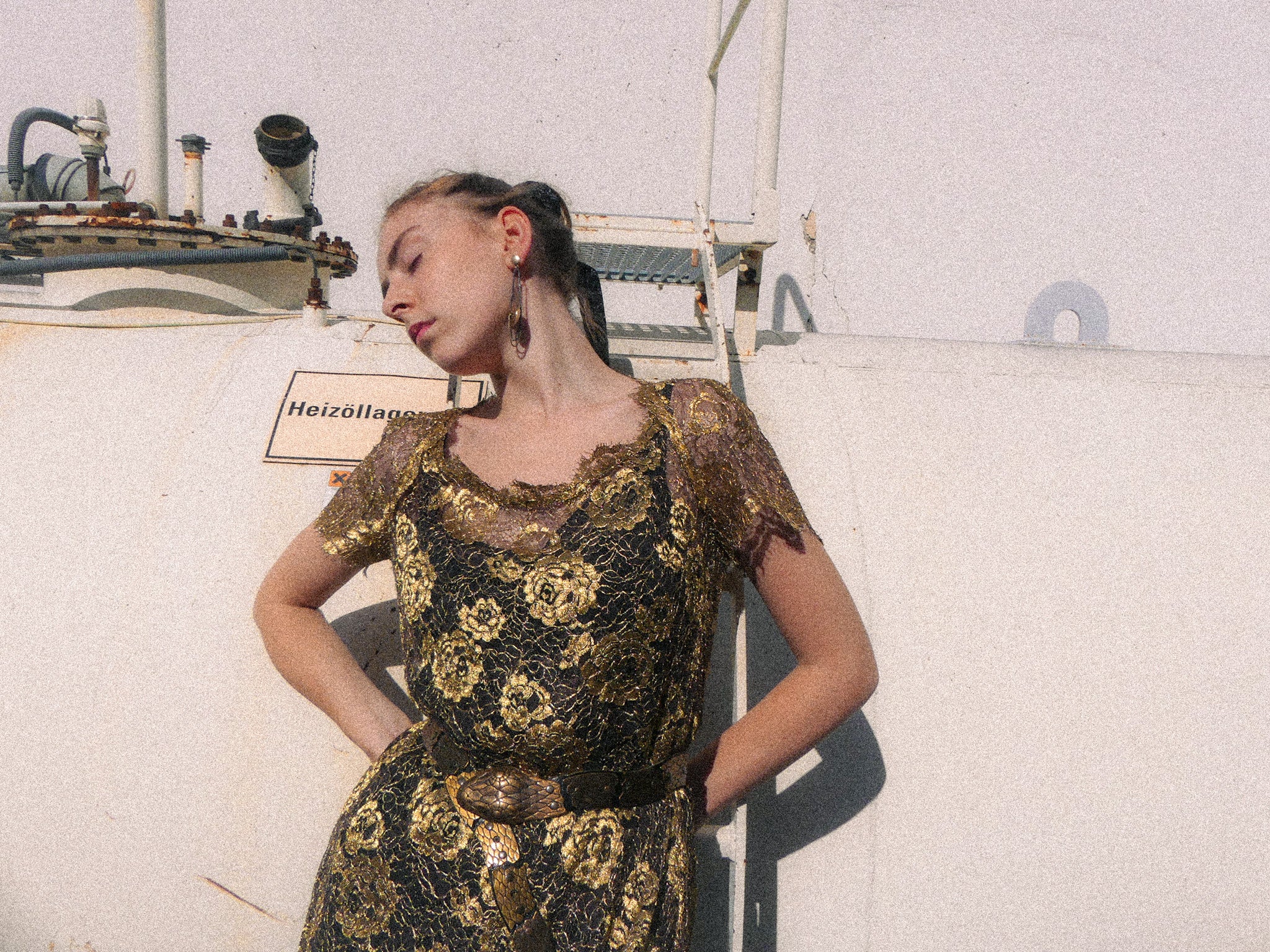 Woman in a black and gold dress standing against a white wall with industrial equipment.