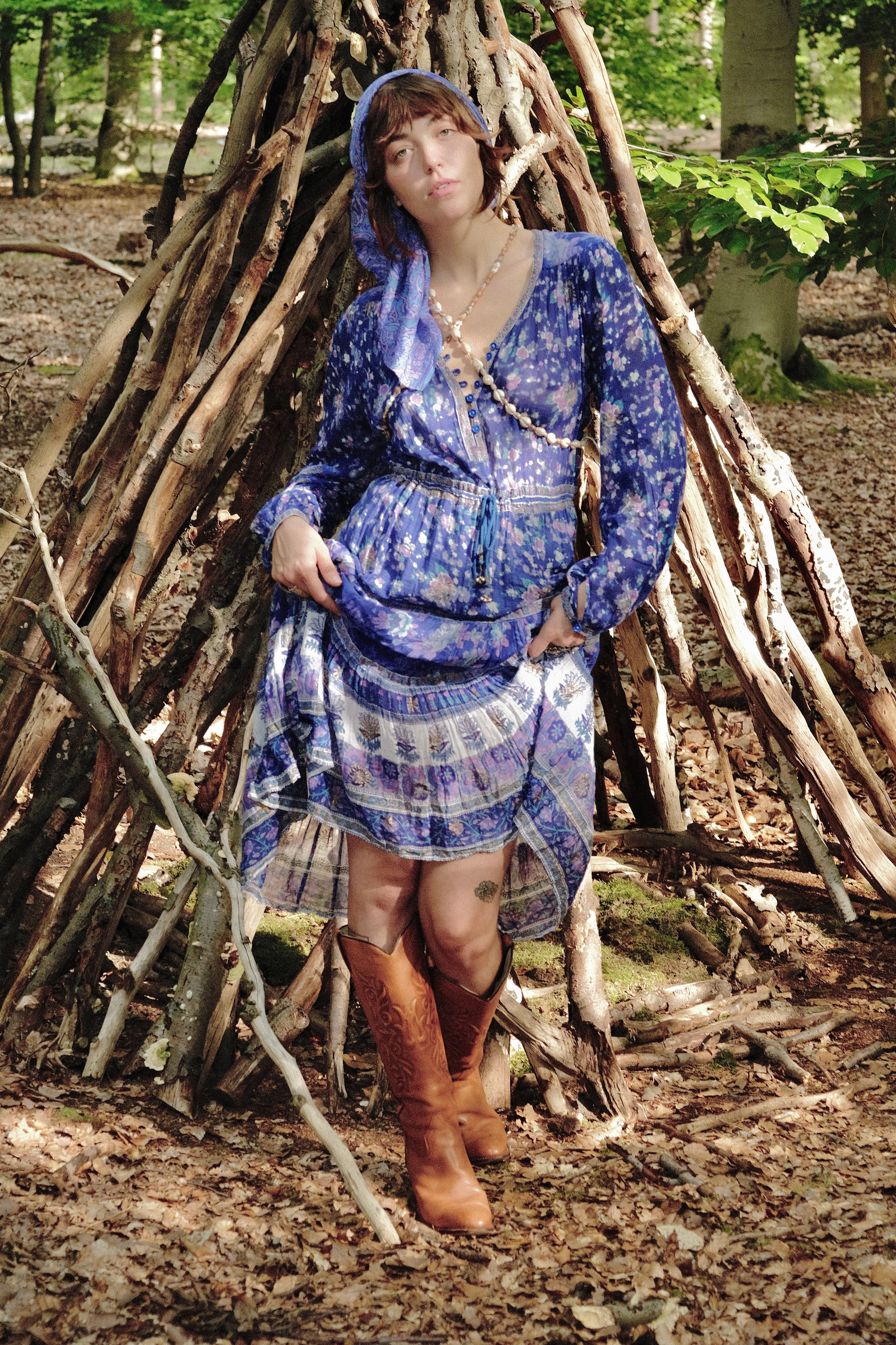 Person in a blue patterned dress standing in front of a wooden shelter in a forest.