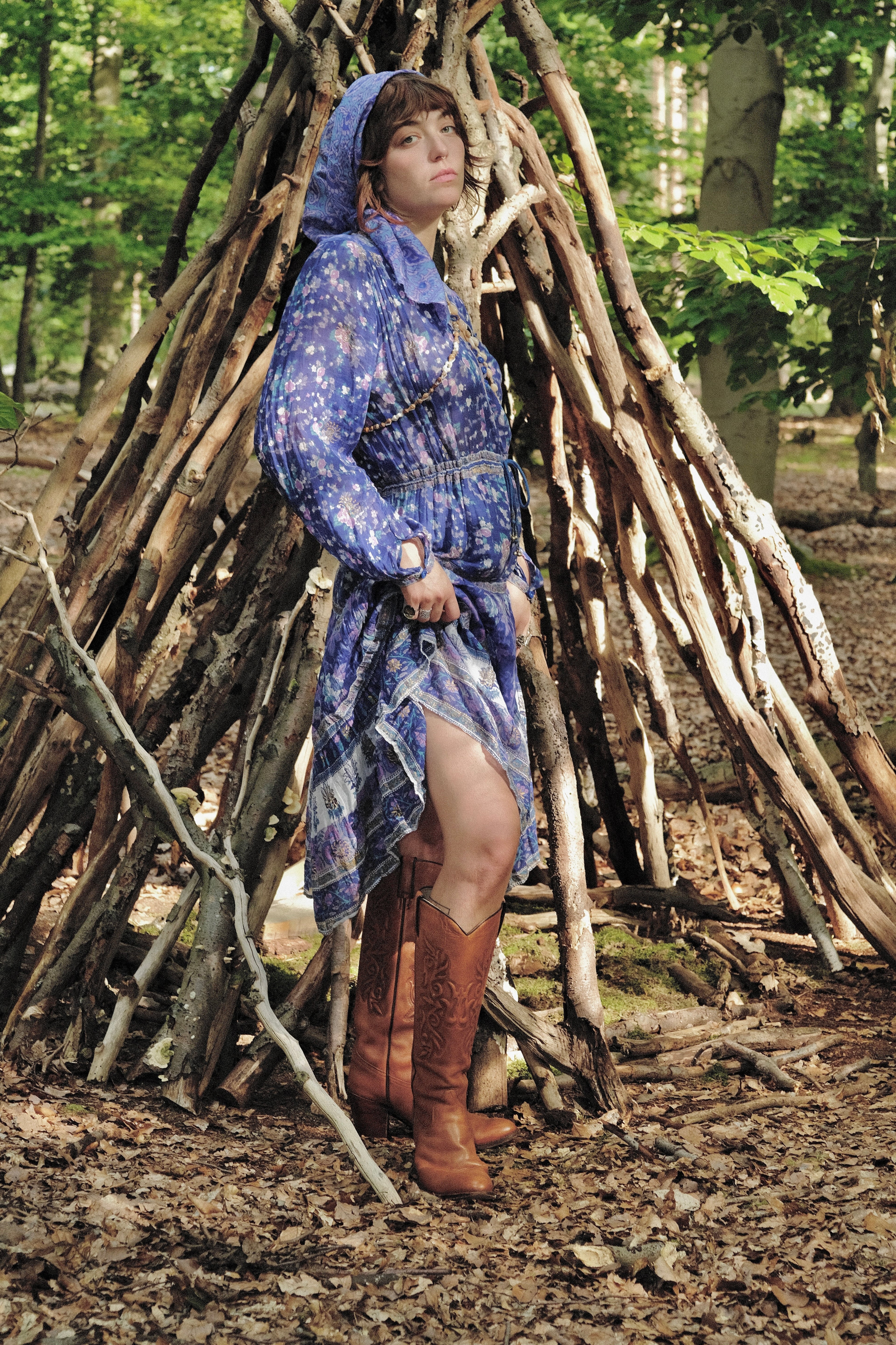 Person in a blue patterned dress standing in front of a wooden shelter in a forest.