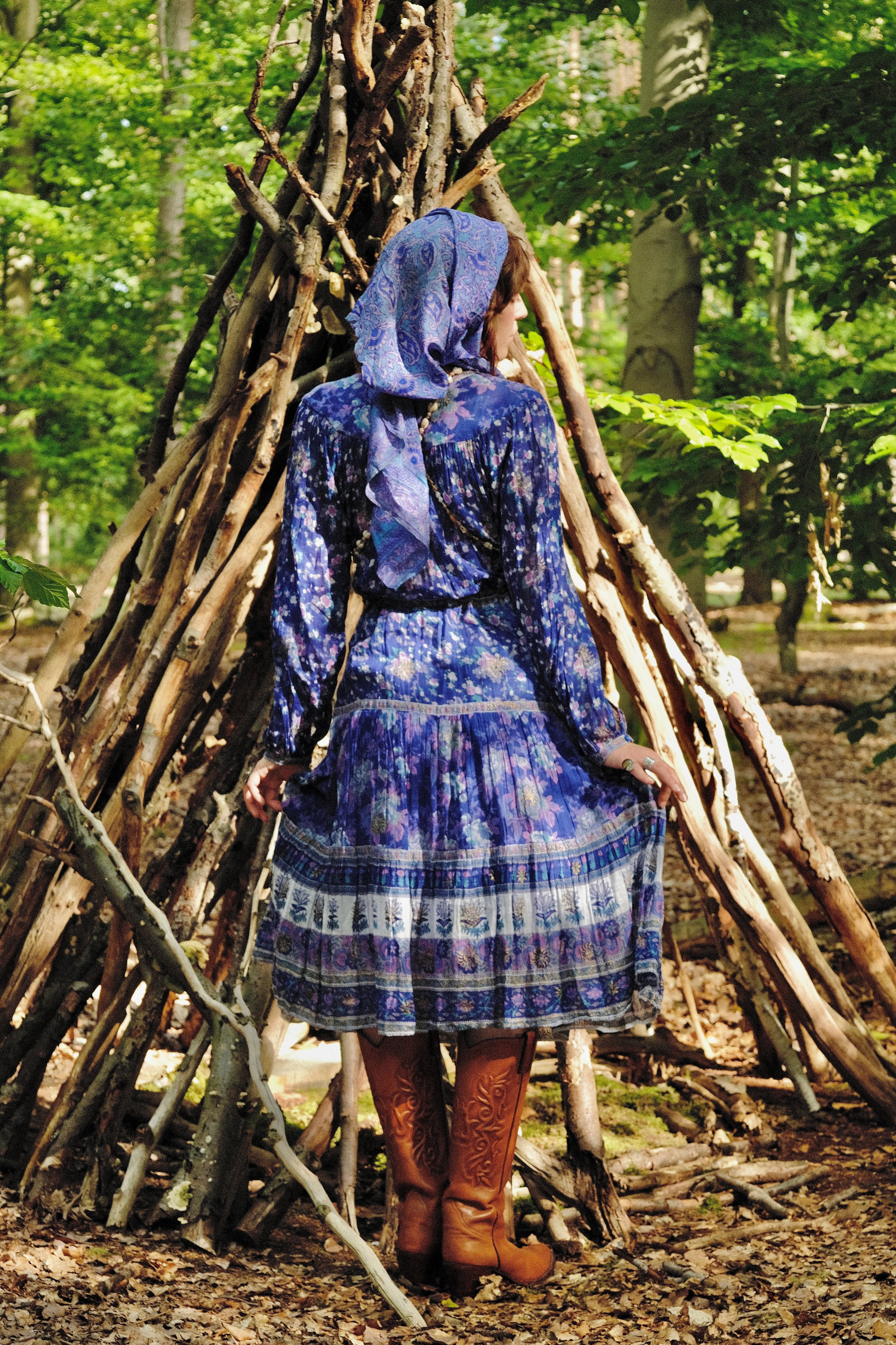 Person in a blue dress standing in front of a wooden teepee in a forest