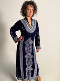 Woman wearing a long navy velvet Moroccan caftan with white embroidery against a plain background