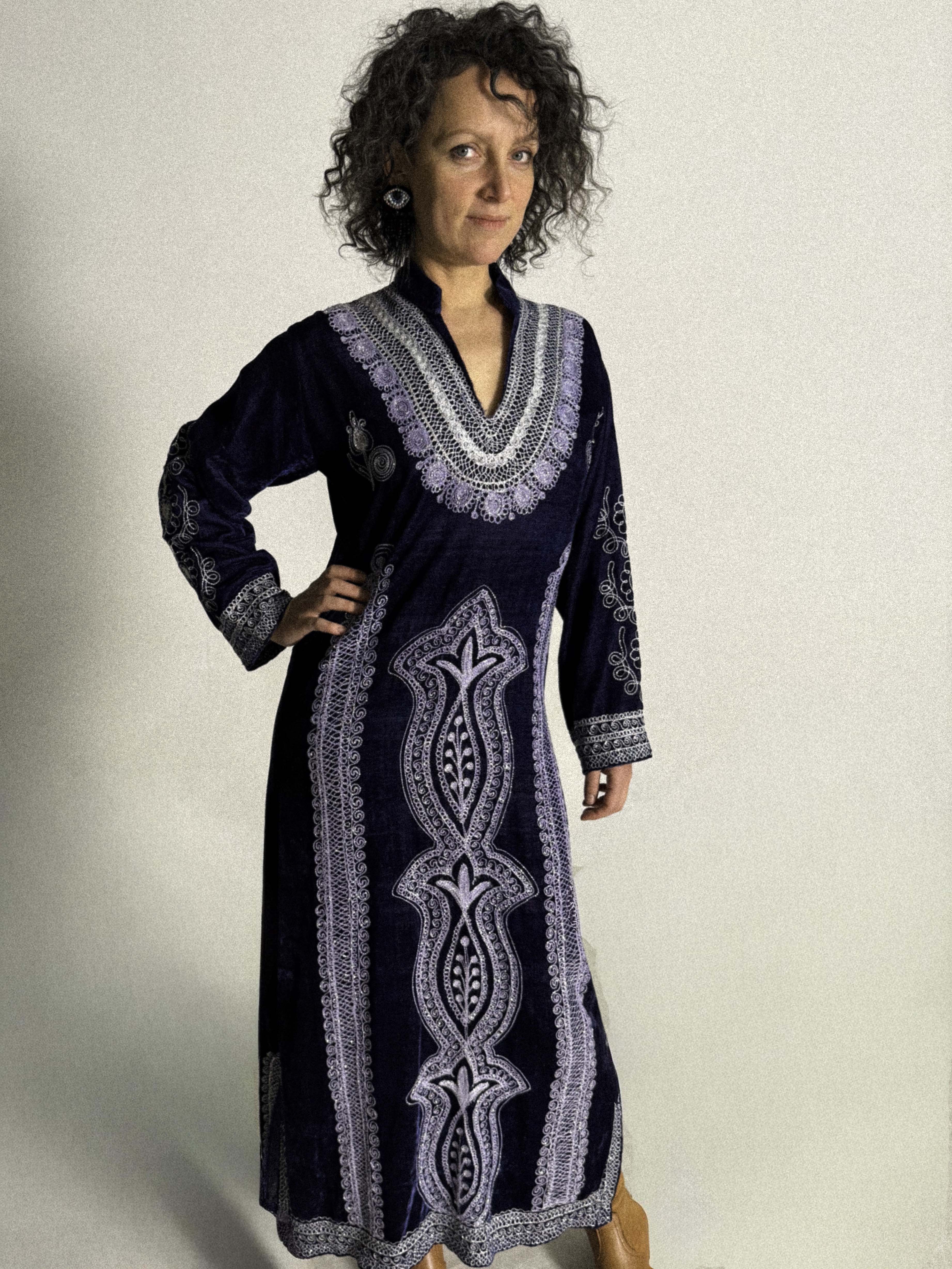 Woman wearing a long navy velvet Moroccan caftan with white embroidery against a plain background