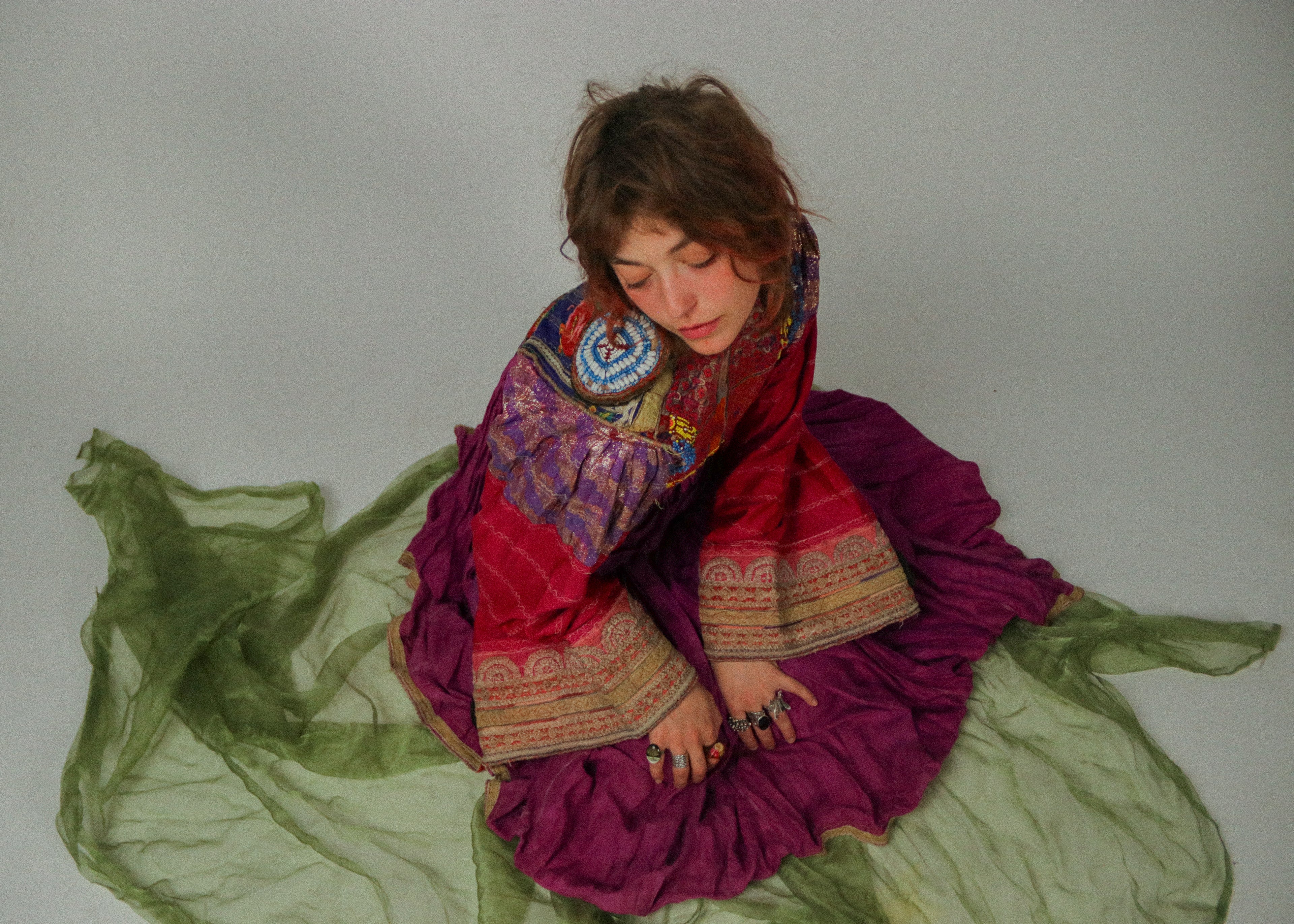 Woman in a colorful traditional afghan kuchi dress sitting on a green fabric against a plain background