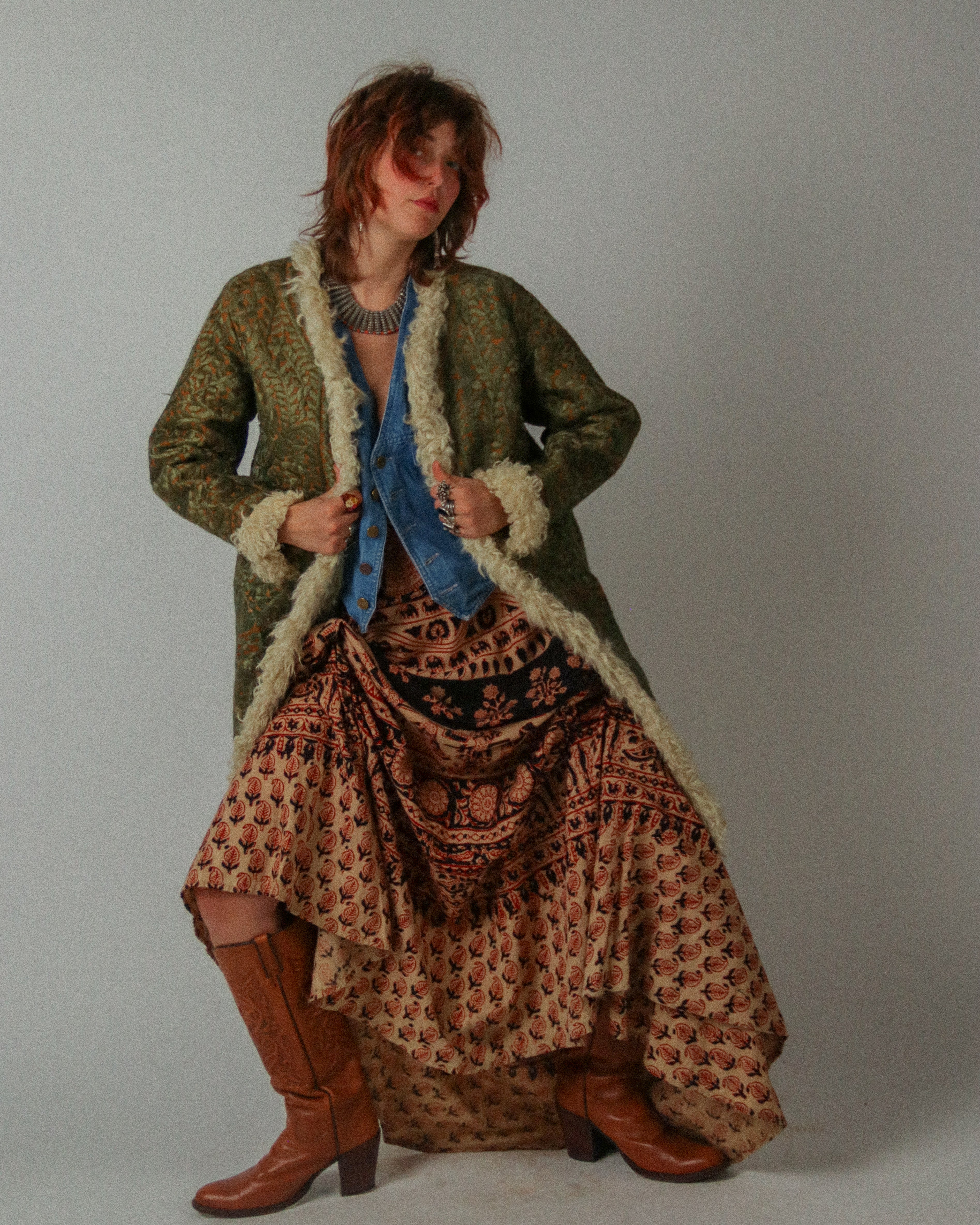 Person wearing a long, patterned coat with a floral skirt and brown boots on a plain background
