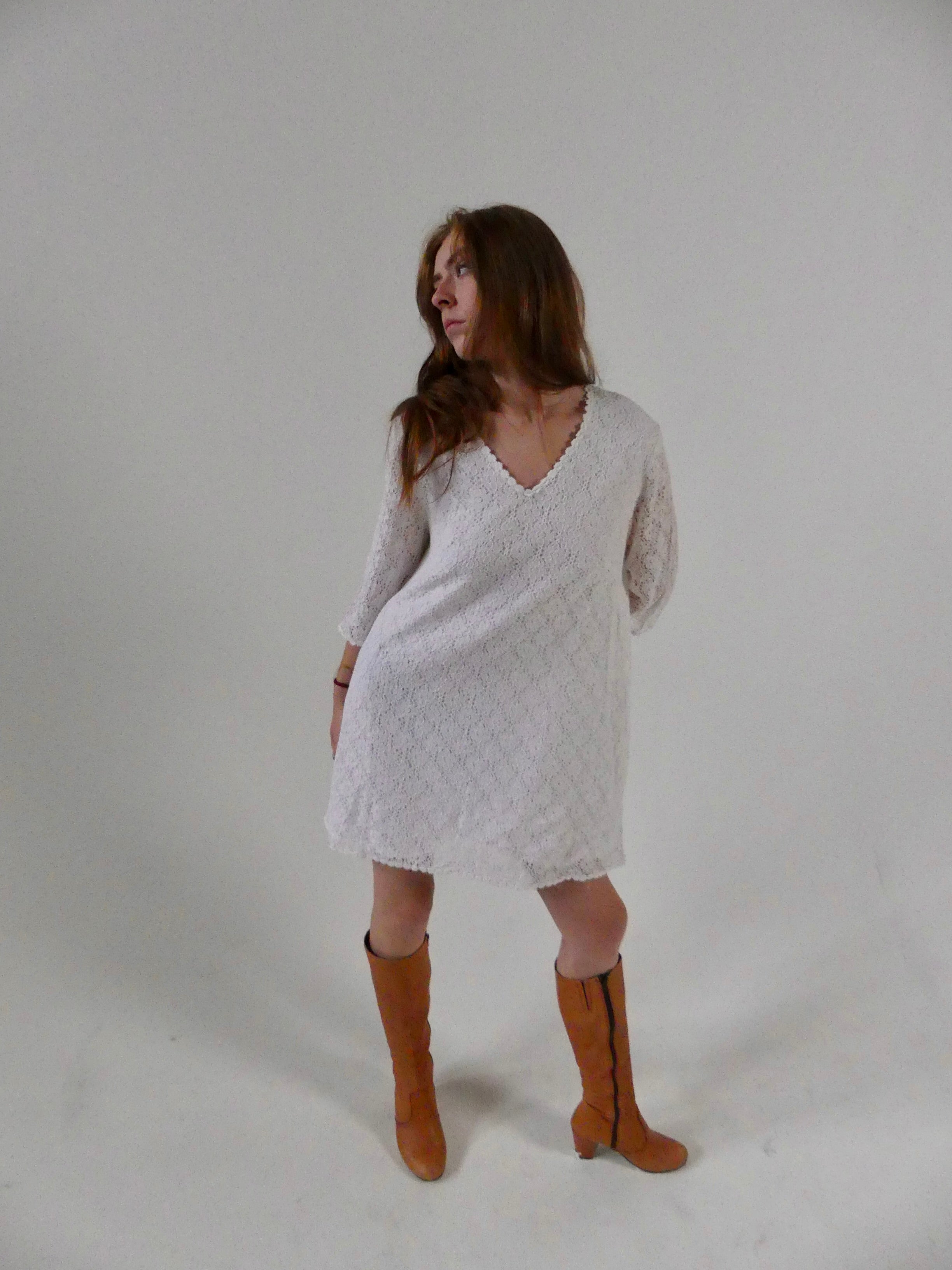 Woman wearing a 1960s White Lace Mini dress with Daisy Trimmings and brown knee-high boots on a plain background